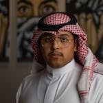 A confident man in traditional Middle Eastern dress and eyeglasses posing indoors.