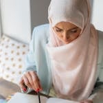 Middle Eastern woman in hijab studying with laptop and notepad indoors.